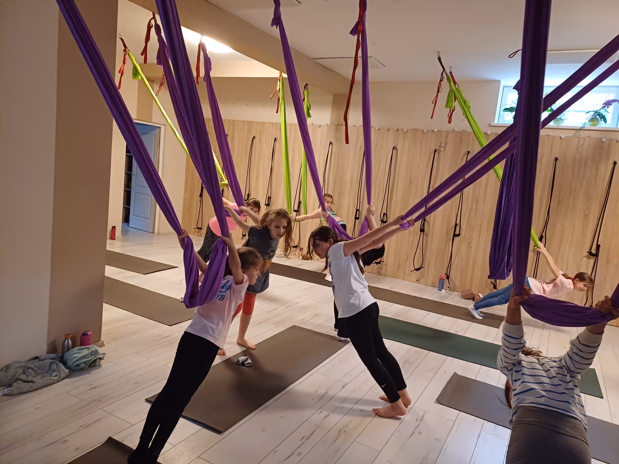 Gyerekek saját testsúlyos jógagyakorlatokat végeznek lila és zöld jógahevederek segítségével egy világos jógateremben.